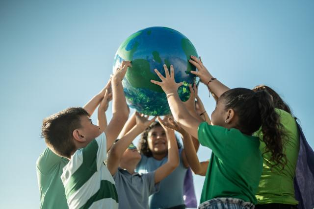 A diverse group of children, outside in green shirts, work together to hold up a ball decorated to look like Earth.