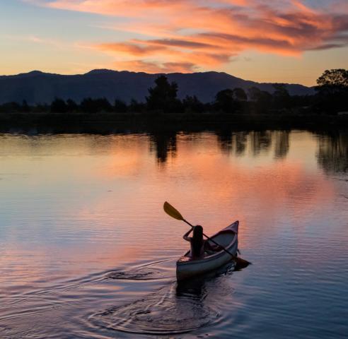 Person canoeing on a lake is silhouetted against the sunset sky.