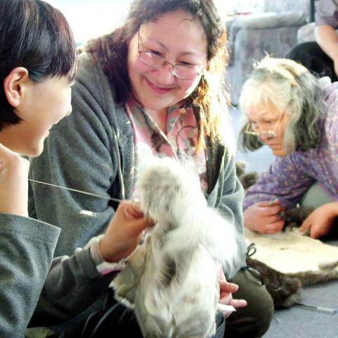 A young woman sews fur while an older woman looks on smiling