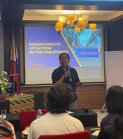 A woman stands in front of a group speaking into a microphone. Behind her a screen displays a PowerPoint slide saying Human rights situations in the Philippines.