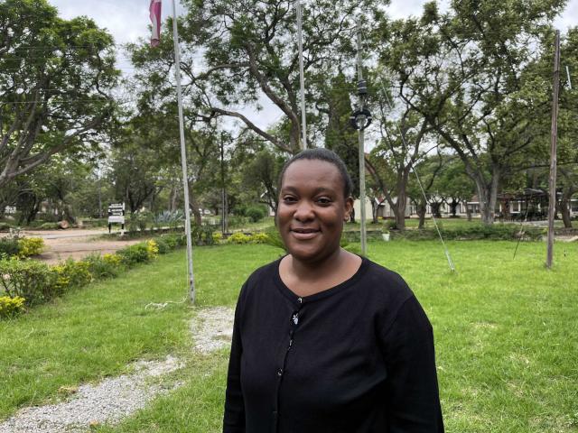 A young woman wearing a black top smiles at the camera against the scene of a lawn and trees.