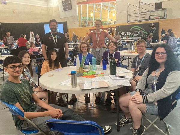 Several young people sit around a table in a gym where a big meeting is happening.