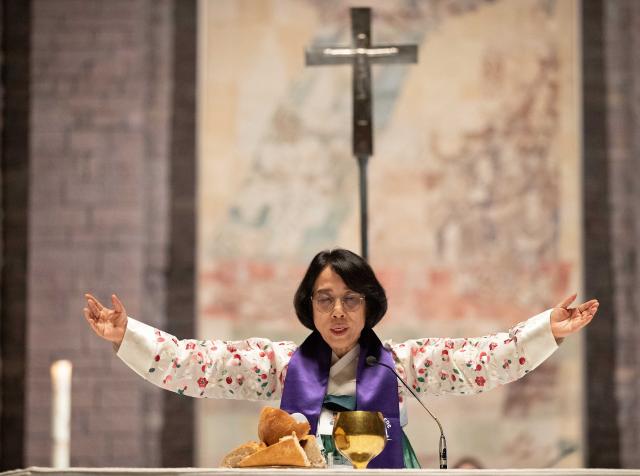 Rev. EunKyung Kim holds her arms out above a communion chalice and bread. A cross hangs on the wall behind her.