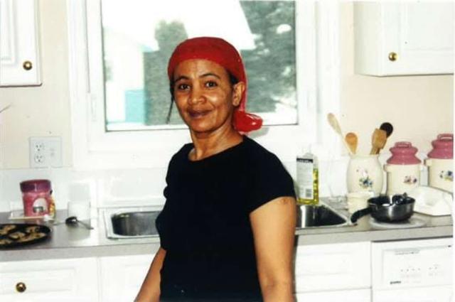 A woman wearing a headscarf stands in her kitchen smiling at the camera.