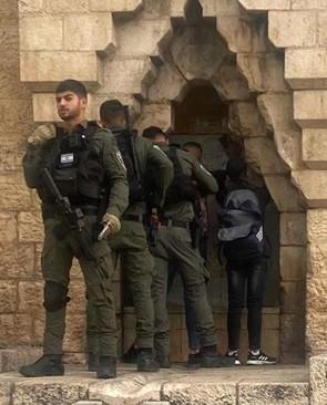 A soldier stands guard in front of a niche in a stone wall where two other soldiers question two teenagers.