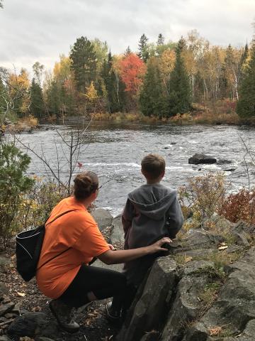 An adult woman crouches down next to a young child as they observe a flowing river, surrounded by autumn trees.