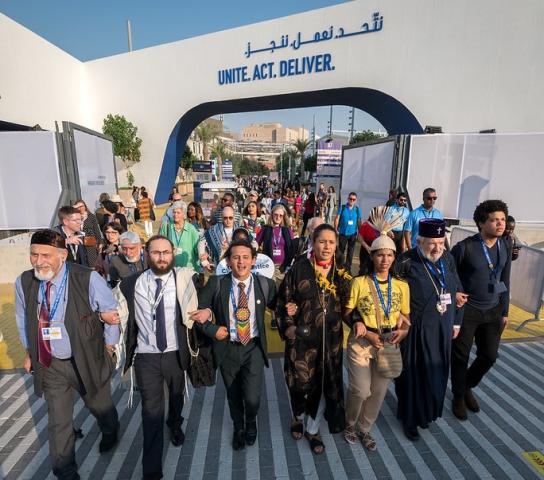 A large group of people walk through an outdoor arch that has Unite. Act. Deliver. printed on it.