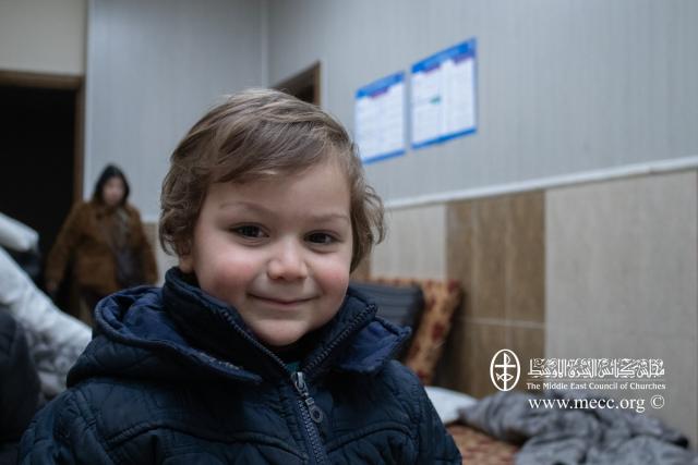 A small boy in a dark blue jacket smiles in a room with mattress on the floor.