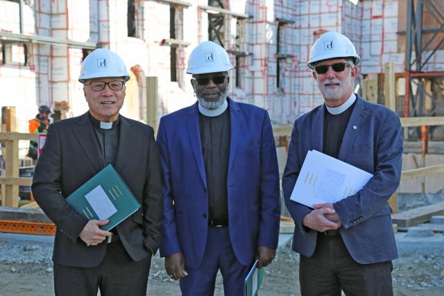 Three ministers wearing clerical collars and hard hats stand in front of a construction site