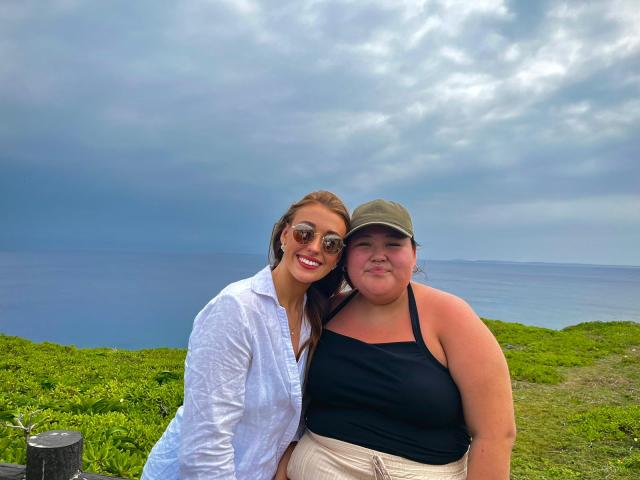 Two young women sit together with the ocean in the background