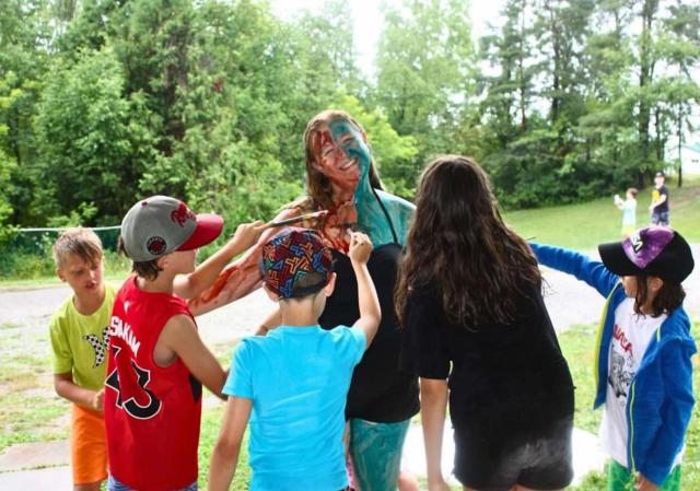 Children surround a camp counsellor with paintbrushes, painting her.