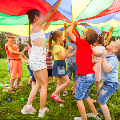 A group of children hold a large rainbow-coloured sail over them.