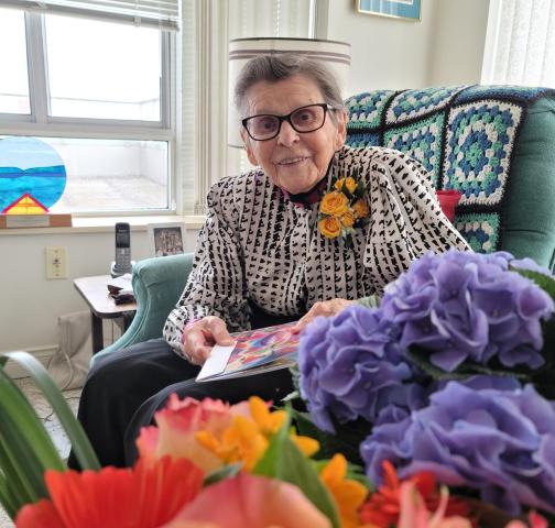 Lois Wilson on her 95th birthday, sitting in big chair and looking at camera. Flowers in foreground.