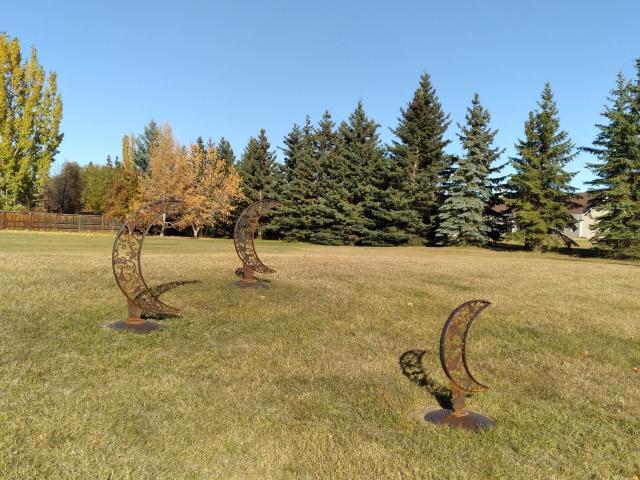 Three metal statues of crescent moons in a field on a nice summer day.