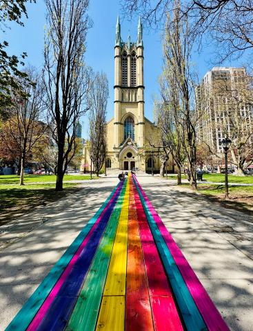 A 100 foot rainbow, pride painted picnic table in front of the cathedral style Met United church