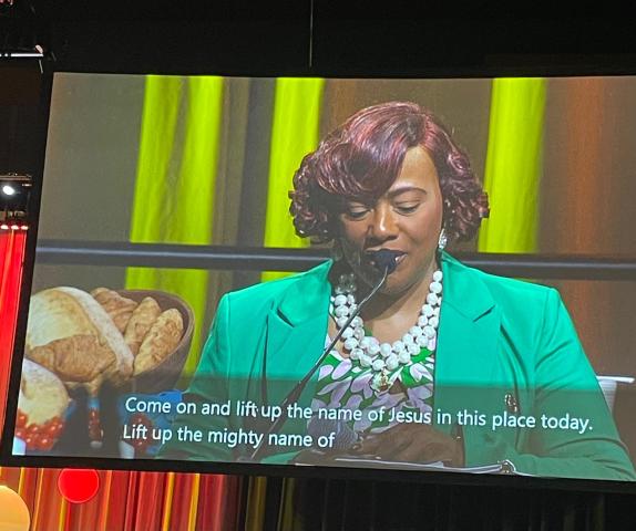 A photo of a woman speaking into a microphone projected onto a large screen