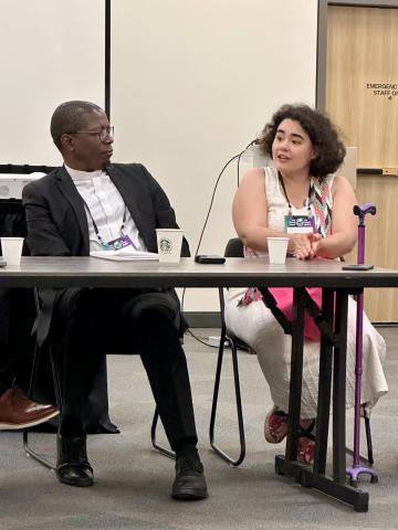 A man and a woman wearing lanyards, seated at a table and speaking.