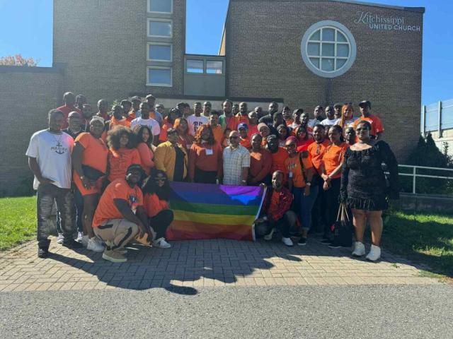 A group of African people, many wearing orange shirts, holding up a pride flag outside of Kitchissippi United Church.