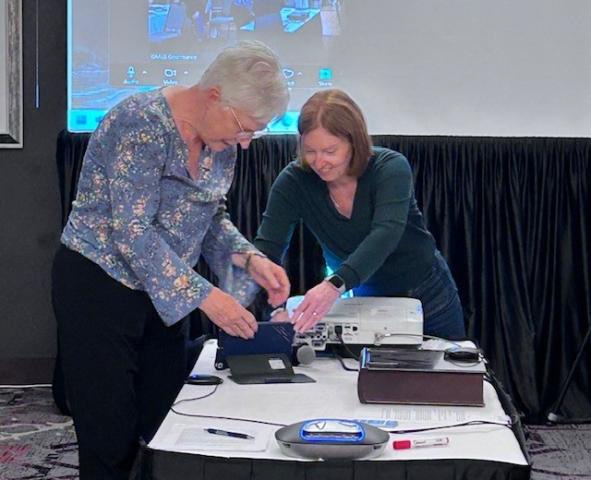 Two women set up electronic equipment on a boardroom table.