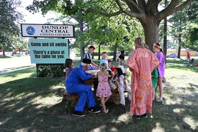 A group of adults and children surrounding a picnic table next to a sign for Dunlop Central United Church.