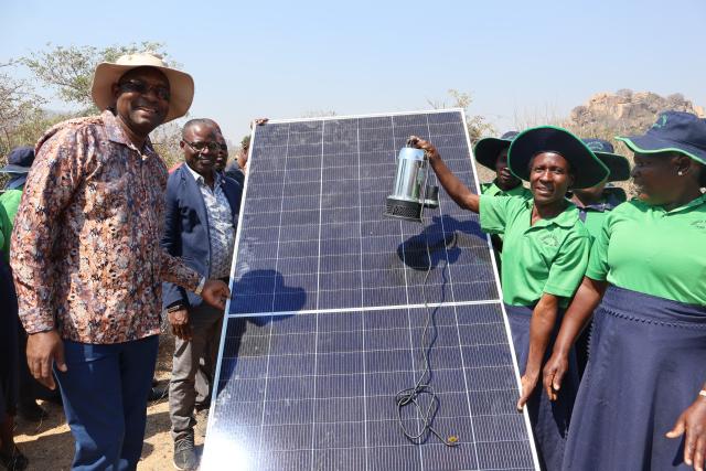 A group of people pose surrounding a solar panel outdoors in Zimbabwe.