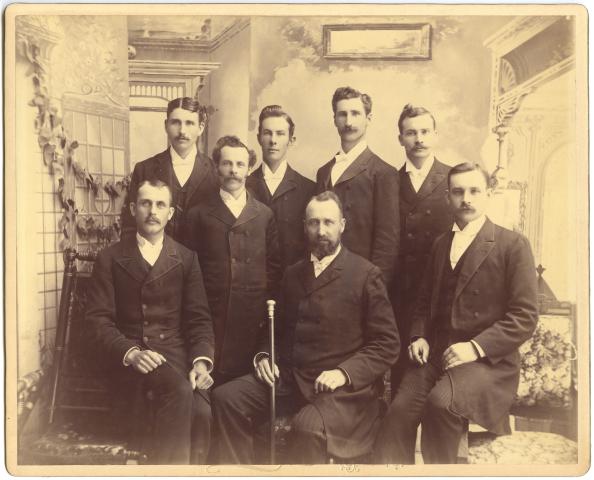 A group of men in 1892, in the formal dress of the time, pose as a group, with the front central figure holding an engraved wooden staff
