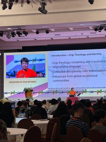 Rev. Dr. Miriam Spies onstage at a conference giving a presentation, with her face projected onscreen behind her.