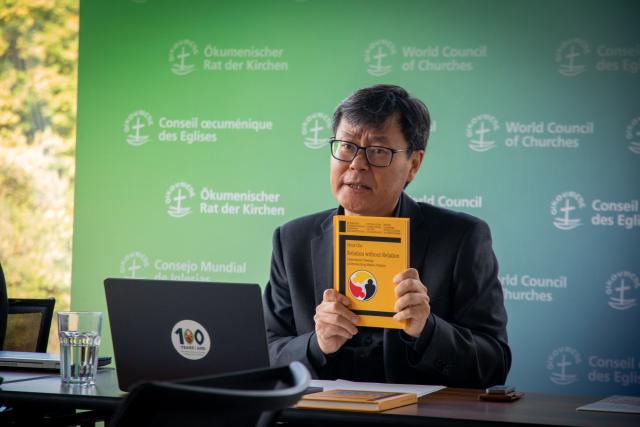 A man in mid-speech holds up a book, sitting down before a laptop with a United Church sticker on it at a desk.