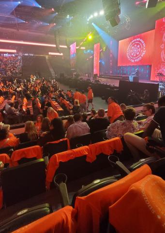 A stadium full of people wearing orange shirts with an orange display on the stage screen, viewed from the right side.