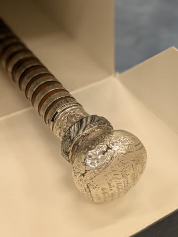 A close-up of the head of a wooden staff, featuring engraved silver panels. 