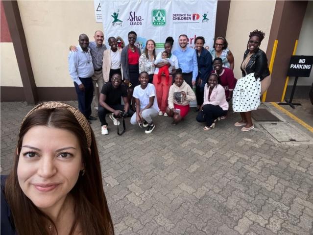 A woman taking a selfie outdoors with a dozen people in the background