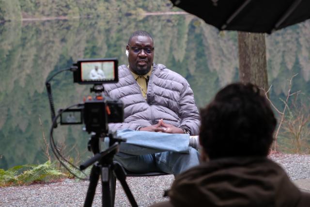 A man being filmed talking on a dock in front of a lake
