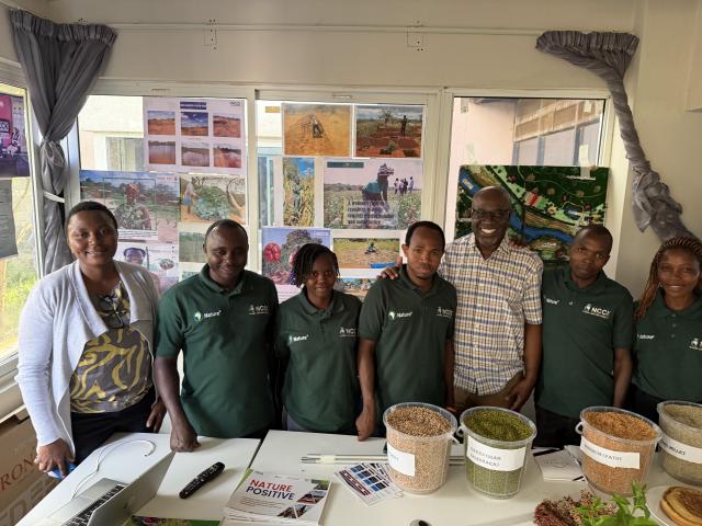 A group of individuals smiling in front of a table holding produce and grains, with photos of farming in action behind them