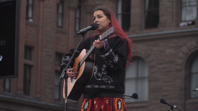 A female Indigenous singer plays an acoustic guitar in front of the Ontario Parliament