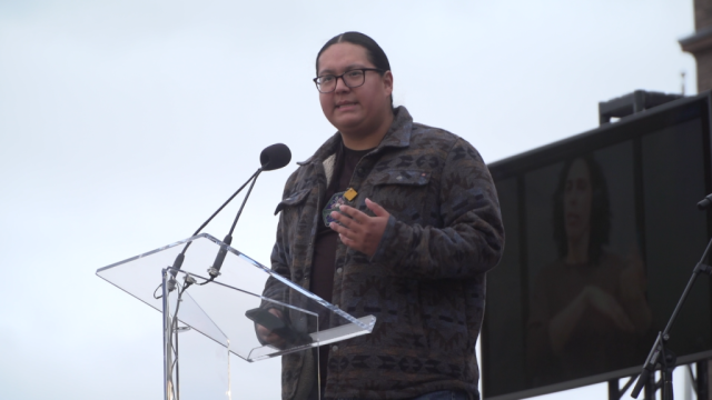 A young Indigenous man speaks onstage in front of the Ontario Parliament