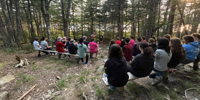 A group of kids sit on benches outdoors in a forest setting