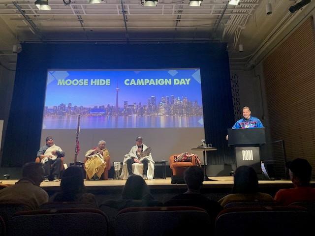 A speaker stands at a podium onstage, in front of a presentation screen that reads "Moose Hide Campaign Day"