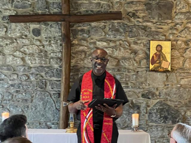 A man preaching in front of a cross and stone wall 