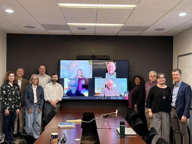 Nine people pose in a boardroom around a tv screen featuring more smiling in an online meeting