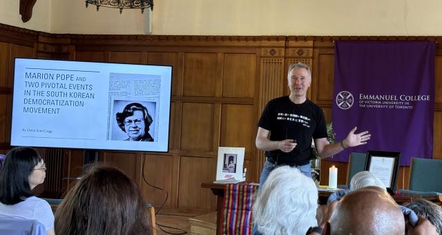 A man gives a presentation in a university room, with an Emmanuel College banner behind him and a photo of Marion Pope onscreen