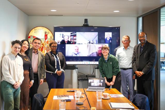 A group of people pose in a boardroom around a monitor featuring faces on a Zoom call