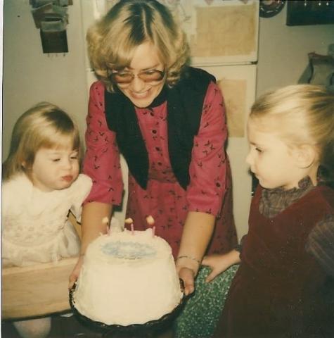 Two young girls celebrate a birthday with cake held by an adult woman in the 1980s