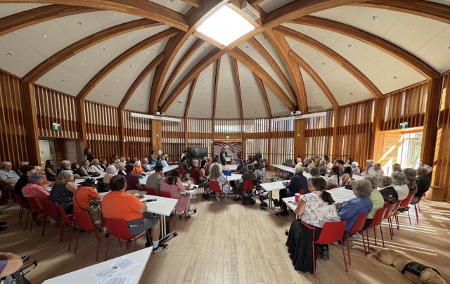 Several dozen people gather at desks in a large room with vaulted timber ceilings.