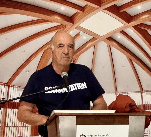 The Very Rev. Dr. Stan McKay speaks at a podium, wearing a shirt that reads "CONSULTATION"