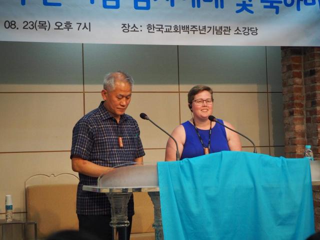 A Korean person and white person speak at a podium with a banner in Korean behind them