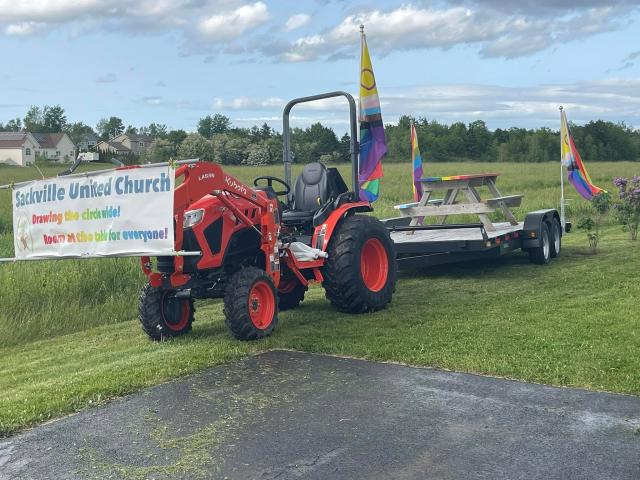 A rainbow picnic table is carried on a trailer by a tractor bearing a "Sackville United Church" banner