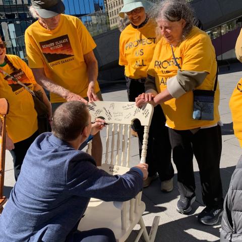 A man crouches down and signs a rocking chair, surrounded by people in yellow shirts