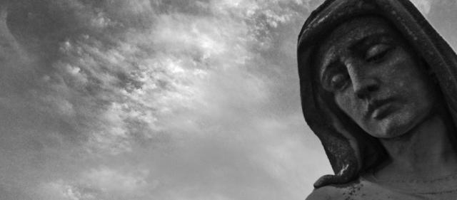 A black and white image of the face of a Mary statue as she looks out beneath a cloudy day.