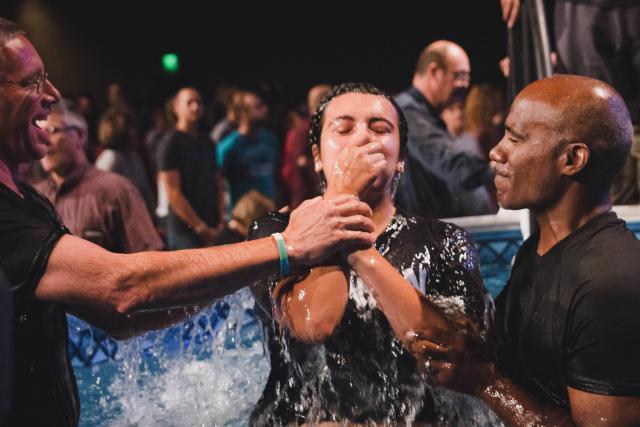 A diverse group of leaders pull a young person holding his nose out of the baptism waters. A number of people in the background await their turn to be baptized.
