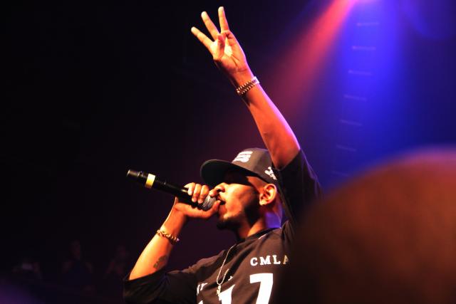 Rapper Kendrick Lamar performs on stage, wearing a black t-shirt and a black baseball cap, he holds a microphone with one hand and raises the other above this head, with three fingers in the air.